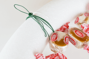 Close-up of a DIY holiday wreath in progress, made with a white foam ring, green wire for hanging, and Goetze’s Caramel Creams® candies attached around the edge.